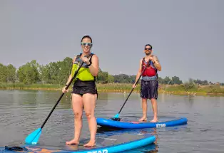 Two happy stand-up paddleboarding students stand up on their blue paddleboards. They are both in swimming attire and are wearing safety vests and sunglasses with paddles in their hands. The water they are on is calm with small rippling waves. In the background is a grassy area with trees and a dusty blue sky.
