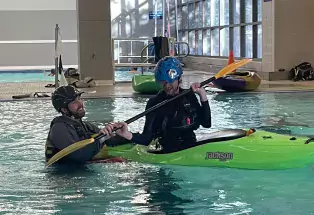 A student sits in a green kayak inside an indoor pool. They are wearing protective gear and are holding a double-sided paddle. Their instructor is in the water next to them. They are both smiling. In the background is another pool illuminated by tall floor-to-ceiling windows letting sunlight beam in.