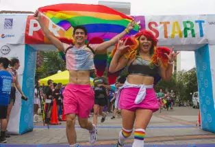 Two joyous people wearing rainbow wings on their backs run across the finish line of the Denver PrideFest 5K race. One of them holds a pride flag tapestry to wave behind them proudly and the other has a rainbow wig on and makes the "I love you" sign with their hands.