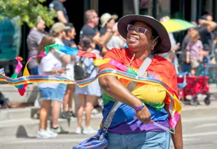 In the foreground is a person in glasses, a bucket hat, a rainbow t-shirt and jeans with a bag across their body. They are smiling peacefully with their eyes closed as they wave thin, long rainbow ribbons on sticks in front of them. In the background is a small crowd of spectators with hats, wagons and other apparel. The sun is shining.