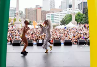 The photographer stands at the back of a large stage in Civic Center Park in downtown Denver. In the foreground are two people dancing on stage singing to each other. One performer is wearing an all-white outfit, a long white wig, and golden pointy boots. The other person is wearing golden underwear and black tennis shoes with a crown on their head. In the background are the tall buildings of downtown and a large audience filled with people and tents.