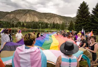 In the foreground are many Mountain Pride celebrators next to a small lake, dressed in various pride flags, including the trans flag, asexual flag, nonbinary flag, lesbian flag, and more. In the middle of the photo is a large rainbow pride flag held by the people surrounding it. In the background are the mountains, trees, and hotels of Avon.