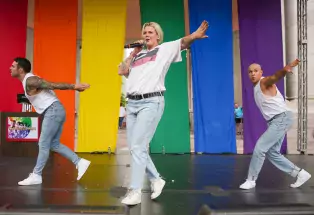 Betty Who, dressed in jeans, a white Nike Air shirt, and white Nike Air sneakers, is in the middle of the photo, singing and performing with an amused expression on a stage in the middle of Civic Center Park in downtown Denver. On the right and left are two background dancers dressed similarly and waving their hands and arms, stuck in dance. In the background are rainbow curtains and Civic Center Park peaking through the cracks.