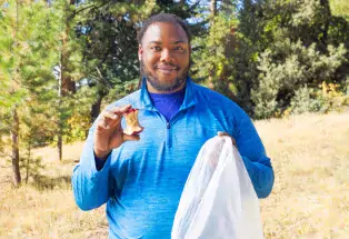A man holds an apple core and a trash bag. He is demonstrating that you need to throw away your eaten apple instead of throwing it on the ground while hiking.