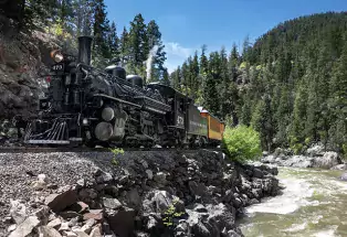 The Durango Train chugs along its track beside a rushing river. Along the sides of the tracks are small cliffs of sharp rocks. In the background is a large hill of pine trees underneath a clear blue sky.