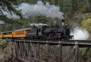 The large Durango Train has steam blowing through the top. The last two cabins in the train visible in the photo are painted pumpkin orange. The sturdy metal tracks that the train is riding along is suspended across a gap between two big rock structures. In the background is a thick grove of dark green pine trees.