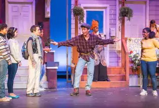 Five students perform on a stage in front of prop houses