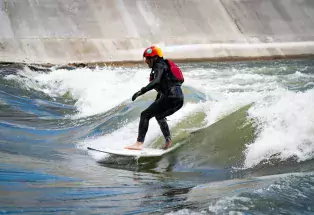 A surfboarder in a black wesuit and red helmet rides waist-high waves between the concret walls of the Pueblo Levee