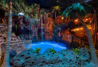 A neon-blue pool is surrounded by fake indoor palm trees and lava with a waterfall flowing into from high above