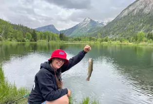 A person holds up a trout they just caught from a shining lake ringed be trees and mountains