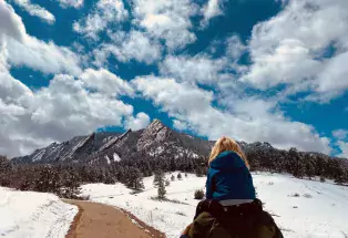 A parent carries a child on their shoulders as they hike a trail lined with snow toward the Flatirons in Boulder, Colorado. The sky is cerulean and is streaked with stark white clouds. 