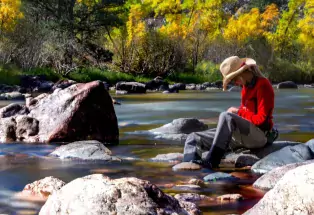 A person in a red sweater, waders and a hat sits on a rock with a fishing rod at the Poudre River.Gardens on Spring Creek in Fort Collins