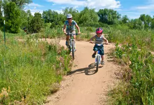 A child and parent ride bikes on a dirt trail surrounded by lush green grass and trees at  Valmont Bike Park in Boulder, Colorado 
