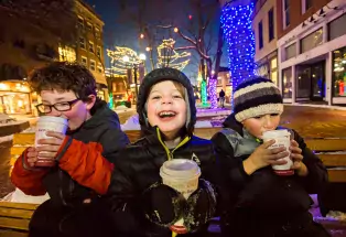 Three children sitting on a park bench on Boulder Colorado's Pearl Street. They're drinking hot cocoa and the trees in the square behind them are decked out in holiday lights.