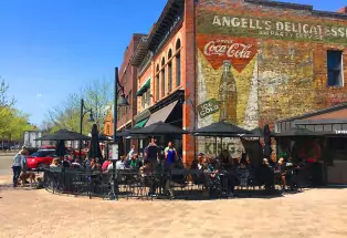 A sunny day on the black-umbrella-covered patio of a large, old-looking brick building that is CooperSmith's Pub & Brewing restaurant in Fort Collins, Colorado. The pati ois packed with people and the sky above is a deep blue. On the side of the building is an old painting of a Coca Cola advertisement.