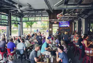 The covered patio of otto PINT in Fort Collins, Colorado is full of sports-watchers as servers walk among the crowded tables on a sunny day. The televisions display advertisements in the background and people on the lawn play games.