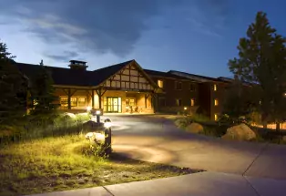A night shot of a short but long cabin lit up by warm outdoor lights sits at the end of a path that curves to the right.  Along the path are plants, pine trees, green grass and two or three short path lights that light up the ground in a circular shape. The sky is a dark blue after sunset.