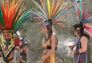 Three Native Americans wearing brightly colored headresses made of brilliant feathers and beautiful beadwork walk with their heads bowed.