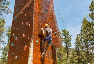 Someone in climbing gear has is scaling a tall dark-red-wood climbing wall outside. The person has both feet secured onto two of the notches and is grabbing two more with both of their hands. In the background is blue sky and a few tall pine trees.