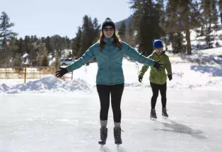 One person stands in the middle of the photograph with their arms stretched out wide to their sides. They are wearing a beanie, sunglasses, winter coat, gloves, leggings and ice skates. They're smiling as they skate along a frozen pond. Another person behind them has their eyes on the ground, trying to focus as they skate. There are pine trees and sunshine in the background.