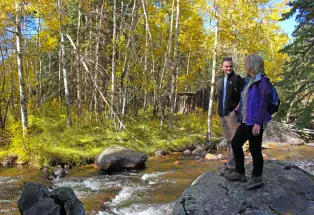 Two smiling hikers clad in outdoor gear stand on a large gray rock. Behind the rock is a flowing river speckled with other smaller rocks and illuminated by the sunlight. Behind the river is a patch of grassy land where a grove of tall, skinny, white aspen trees with yellow leaves sits beneath a blue sky.