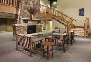 A tall fireplace adorned with stone reaches to the top of the photograph on the righthand side. Around it are several wooden-framed chairs and loveseats with a few dark brown circular tables in the mix. In the background is a grand staircase leading to a second floor.