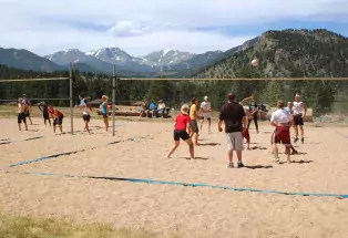 A photograph of a large rectangle of sand. In the sand stands two volleyball nets, although one of them is not fully visible. The boundaries are marked by blue rope in the sand. There are about 30 people in the sand playing volleyball on both courts. Someone hits the ball up in the air and it hangs there as the picture was taken. In the background are the snowcapped Rocky Mountains and other smaller hills covered with trees a little closer to the subjects. The sky is cloudy other than a sliver of blue.