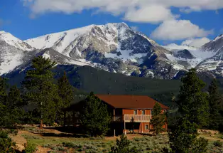 In the foreground is a stout, red, multi-leveled cabin somewhat hidden by sporadically grown dark green pine trees. In the background is a majestic image of the Rocky Mountains covered in snow towering over the cabin. The sky is blue and dotted with fluffy clouds. 