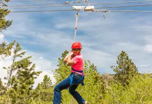 A person strapped into a zipline and gear, including a bright red helmet and sunglasses, smiles widely as they glide through the air. Behind them are an array of green pine trees and wispy clouds in a blue sky.
