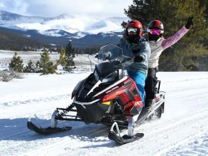 Tour the Leadville and Twin Lakes area by snowmobile. Photo Credit: Instagram/@ingridcg.fit Two people wearing helmets, goggles and winter gear ride a red snowmobile near Leadville with snow-covered mountains in the distance.