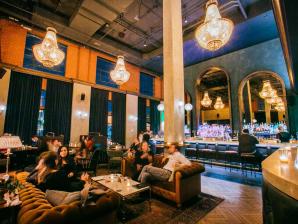 Death & Co at the Ramble Hotel Bar patrons in a cozy, jewell-toned bar relax on plus couches with glowing chandeliers above them