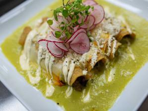 A square white plate holds four enchiladas smothered in green sauce, zigzagged by sour cream and topped with slides of radish